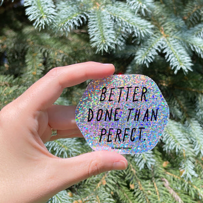 Hand holding a holographic sticker with 'Better Done Than Perfect' text in front of a green tree.