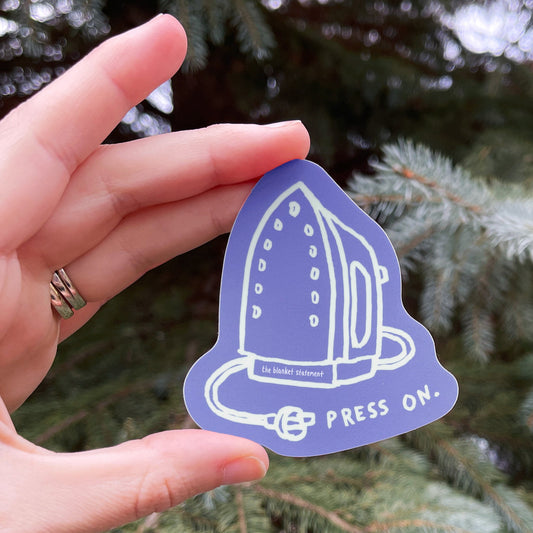Hand holding a perwinkle and white sticker with "Press On" and a doodle of an iron against a green tree background